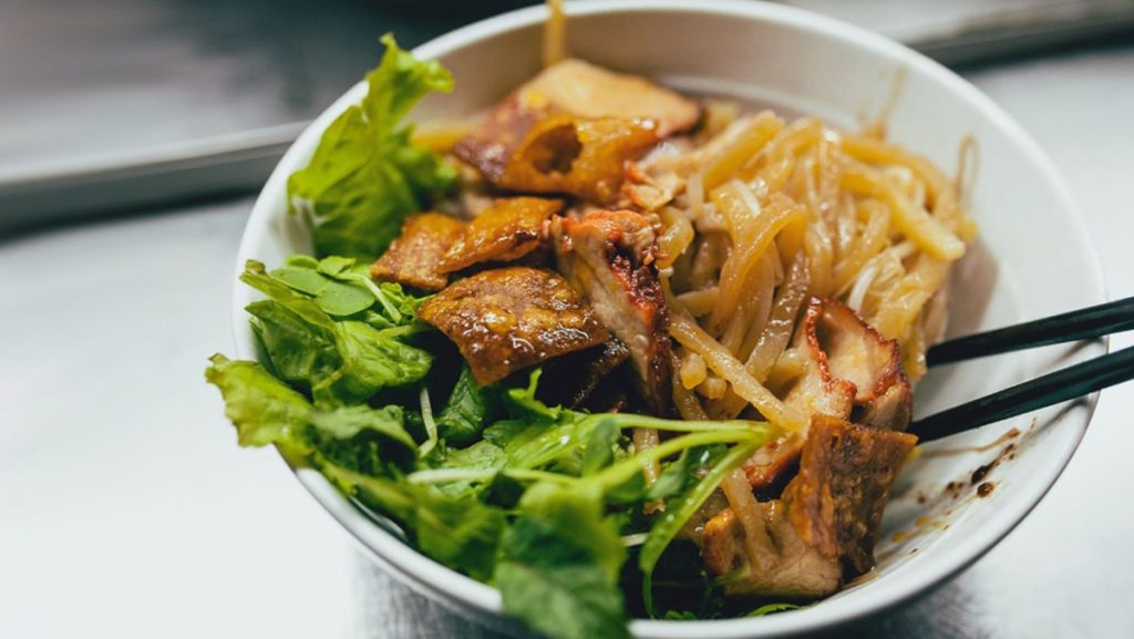 A bowl of Cao Lầu, showcasing thick noodles, slices of pork, fresh herbs, and crispy wonton crackers