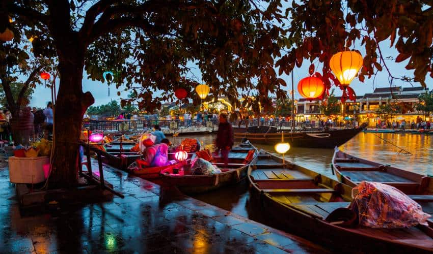 A wooden boat gliding along the Thu Bồn River, with lush greenery and traditional buildings in the background