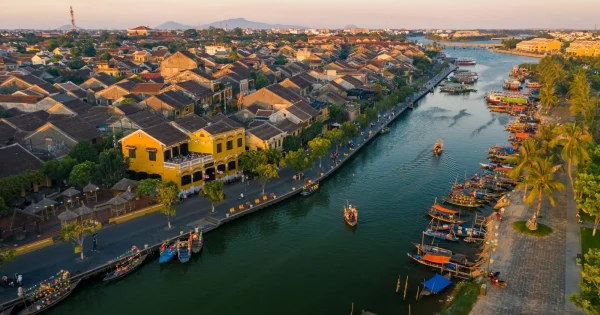 Scenic view of Hoi An in January, with peaceful streets lined with traditional yellow-tinted buildings, lanterns glowing softly in the cool winter evening. The riverside is calm, reflecting the warm hues of sunset, and locals and tourists wander through the charming, historic town under clear blue skies.