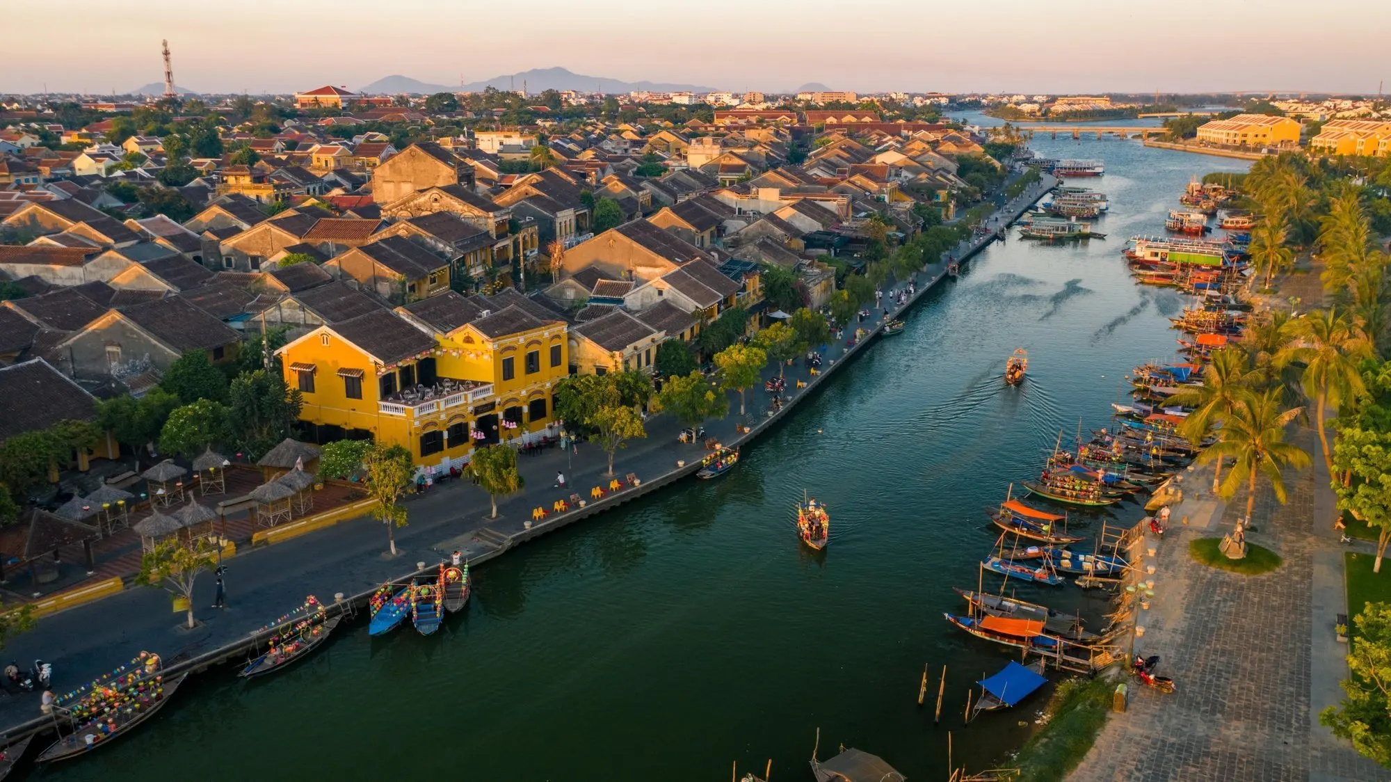 Scenic view of Hoi An in January, with peaceful streets lined with traditional yellow-tinted buildings, lanterns glowing softly in the cool winter evening. The riverside is calm, reflecting the warm hues of sunset, and locals and tourists wander through the charming, historic town under clear blue skies.